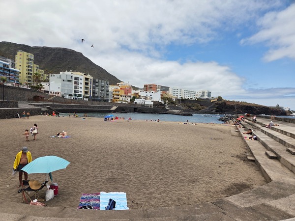 Spiaggia principale di Bajamar con sabbia scura, gradinate e case colorate davanti al molo.