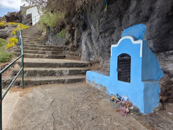 Piccola cappella dipinta di azzurro incastonata nella roccia, con offerte floreali alla base.