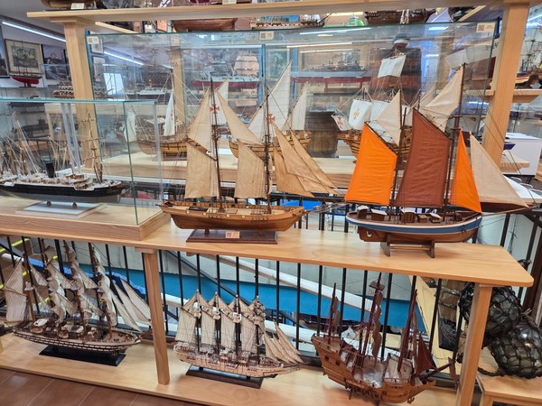 Serie di modelli navali a vela, con una goletta dalle vele arancioni in primo piano esposta su mensole in legno.