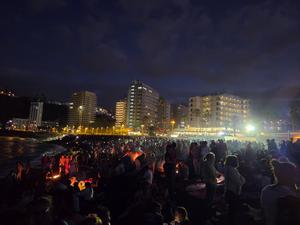 Vista notturna della spiaggia affollata con piccoli falò e persone riunite in cerchio.