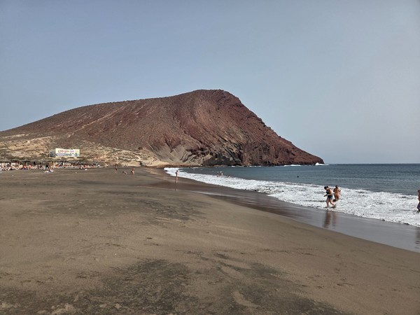 Primo piano della Montaña Roja vista dalla battigia di La Tejita con bagnanti e sabbia bagnata in primo piano.