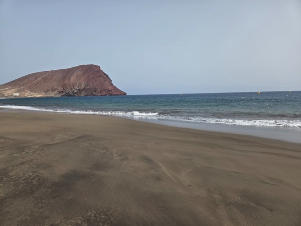 Panoramica ampia della spiaggia di La Tejita con litorale curvo, acqua turchese e zone attrezzate visibili sulla sabbia scura.